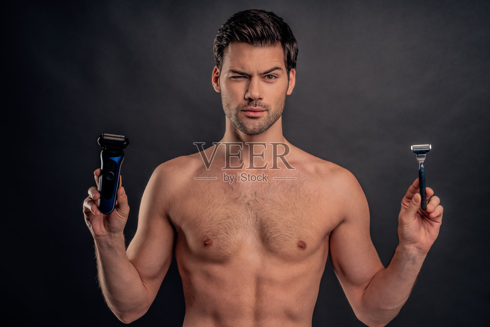 英俊的留着胡子的年轻人孤立无援。赤裸上身的肌肉男子的肖像是站在灰色的背景与修剪在一只手和剃须刀在另一只手。男性保健的概念。照片摄影图片