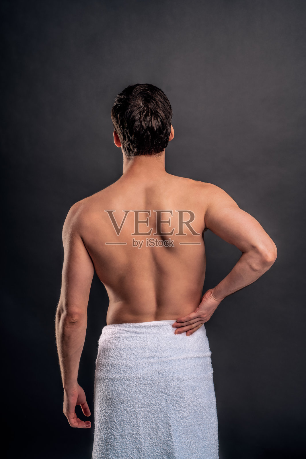 英俊的留着胡子的年轻人孤立无援。赤裸上身的肌肉男子的肖像是站在灰色的背景。男人拖着他的后腿。经历背痛。照片摄影图片