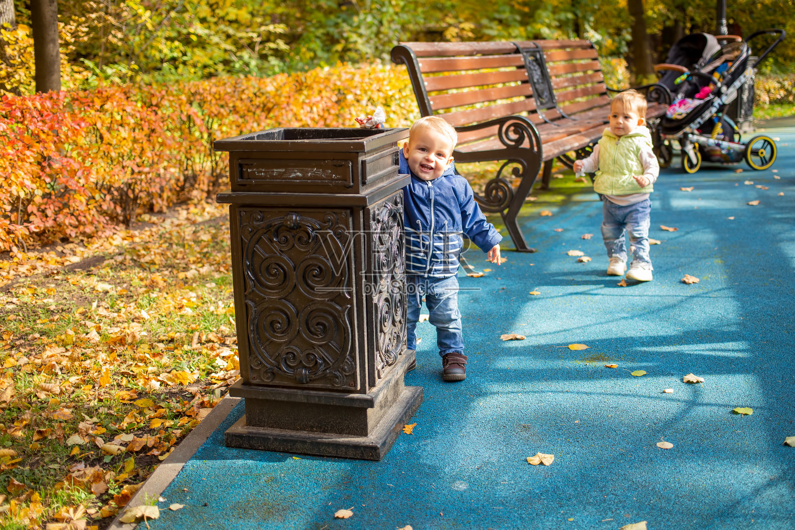 一个可爱的小婴儿穿着蓝色的衣服站在一个公园的街边瓮旁，在那里扔垃圾，微笑着看着镜头。另一个孩子在后面。软焦点，背景模糊。照片摄影图片