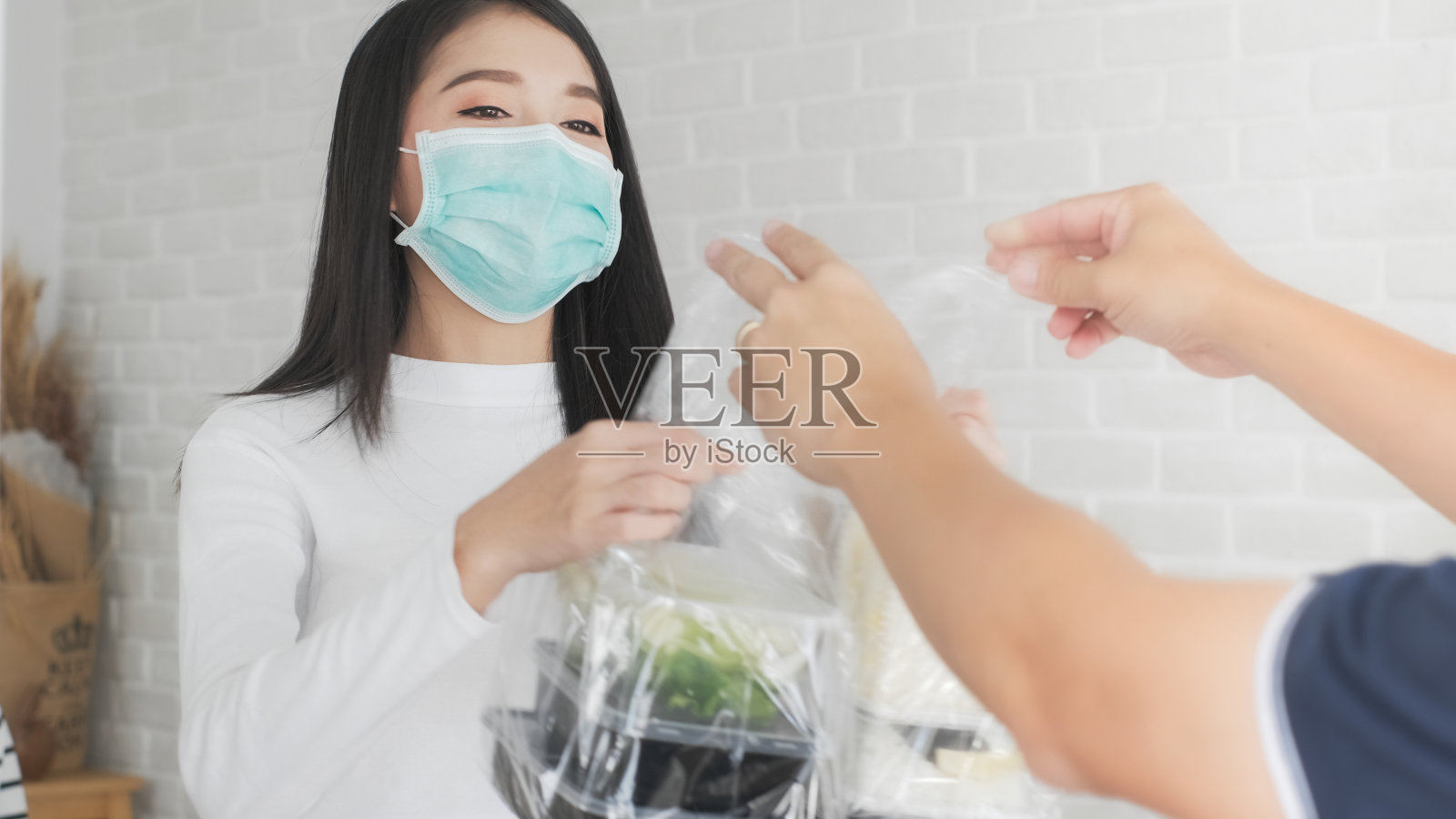 亚洲妇女在家里戴口罩，并将戴口罩的男子处理袋装的食物，水果，蔬菜在家门口送给女性顾客。2019冠状病毒病期间的杂货配送服务。照片摄影图片