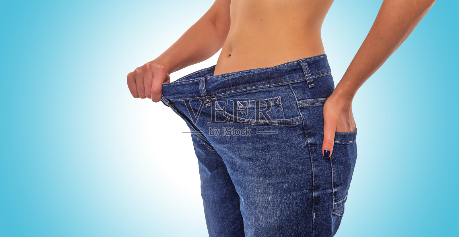蓝色背景下穿着大牛仔裤的苗条女孩。减肥。照片摄影图片