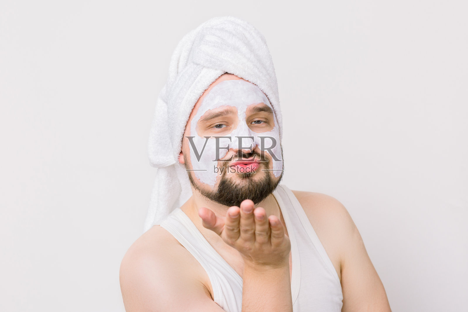 情绪幽默的男人早上洗完澡后戴着白色的清洁面膜，看着镜头，做了一个飞吻。近距离的肖像，孤立的白色照片摄影图片