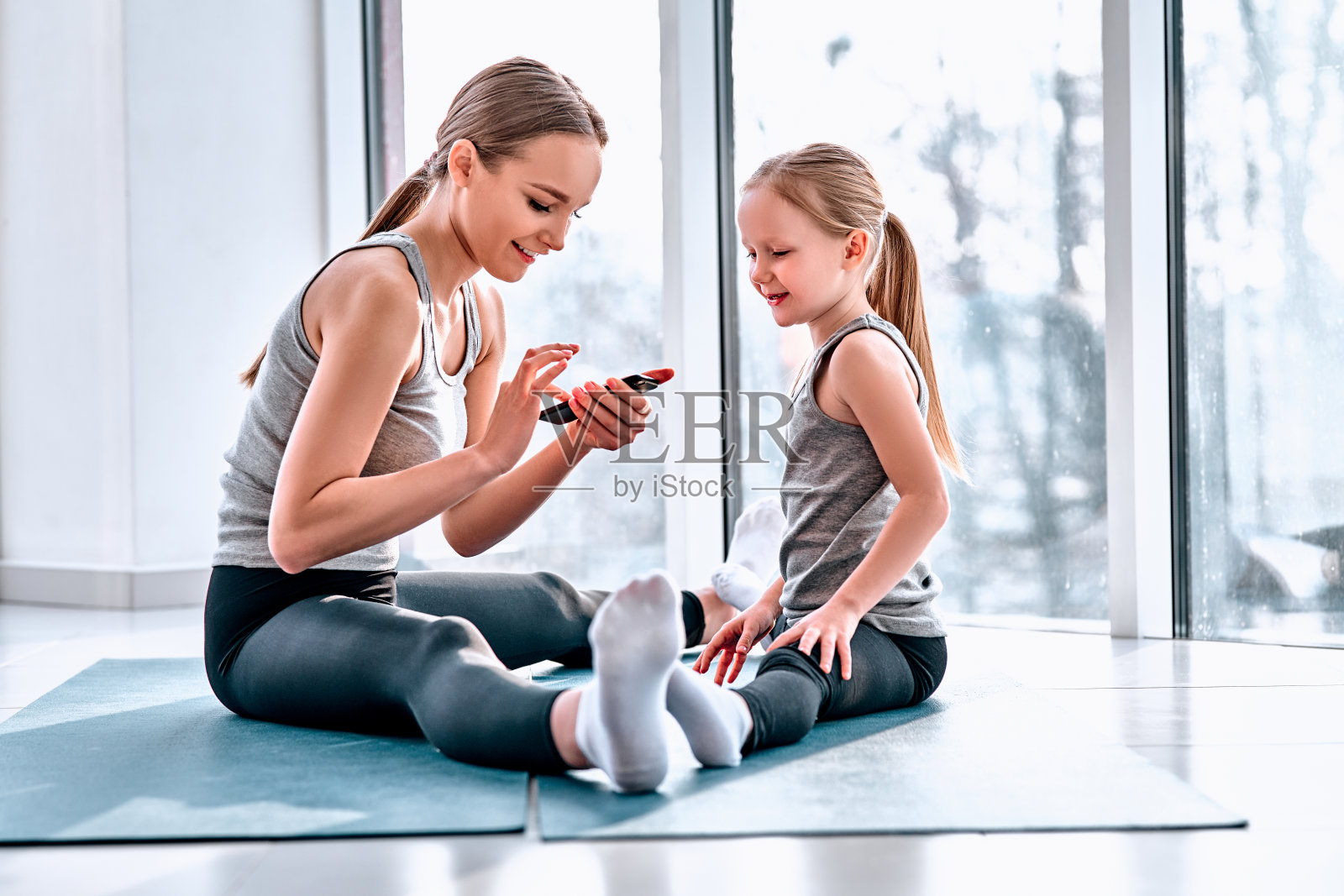妈妈和女儿在做瑜伽。一家人在健身房。小女孩和穿着白色t恤和黑色打底裤的妈妈在一起。女孩们坐在绿色的垫子上照片摄影图片