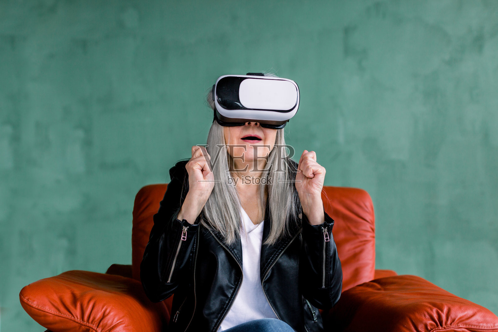 工作室拍摄的令人惊讶的漂亮的高级女人灰色头发，穿着时尚的皮夹克，坐在红色的软扶手椅上特殊的3d虚拟眼镜，保持她的拳头紧握照片摄影图片