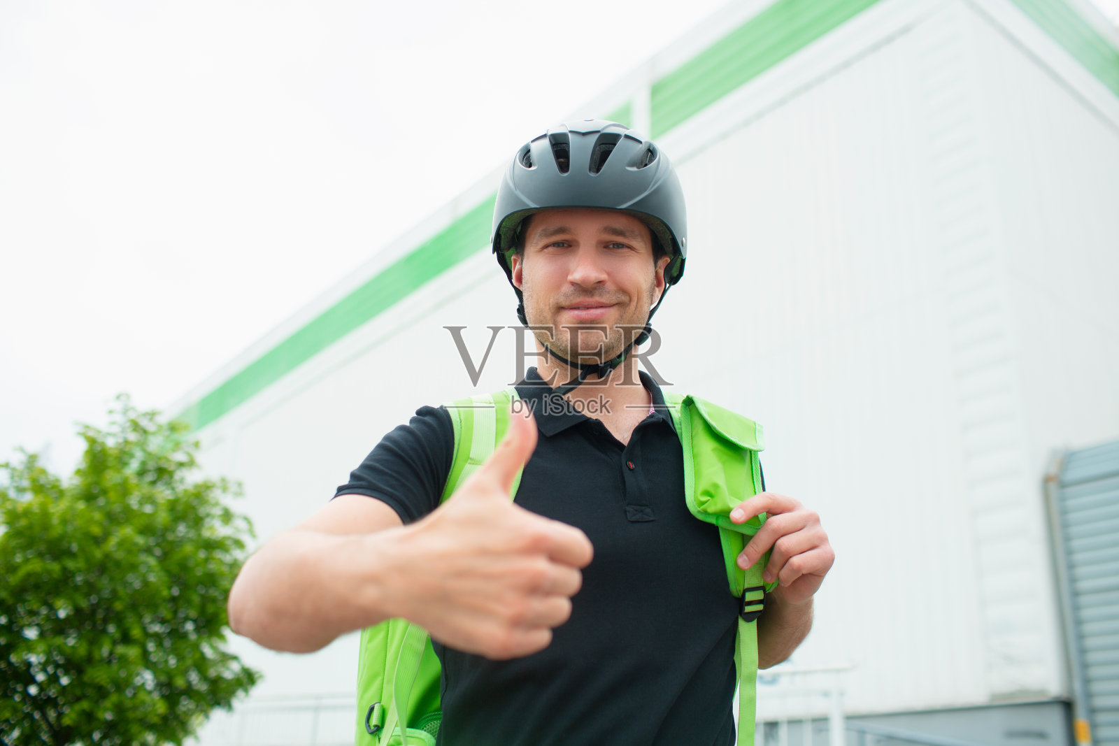 食品配送的概念。送餐员用智能手机更快地联系顾客。快递员有一个装在绿色背包里的冰箱。他看着摄像机，竖起大拇指。照片摄影图片