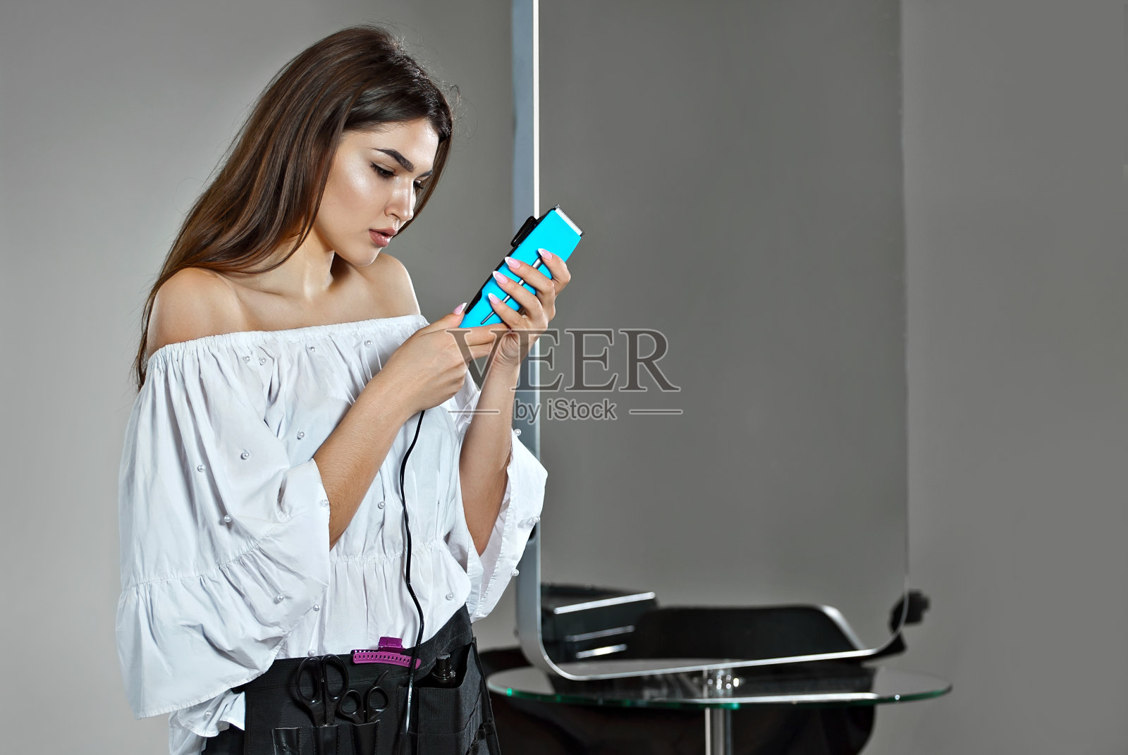 美丽的女孩发型师在一个灰色的背景。穿着白衬衫的年轻女孩，手里拿着剪刀。在模型附近放置文本。布局设计和复制空间。照片摄影图片