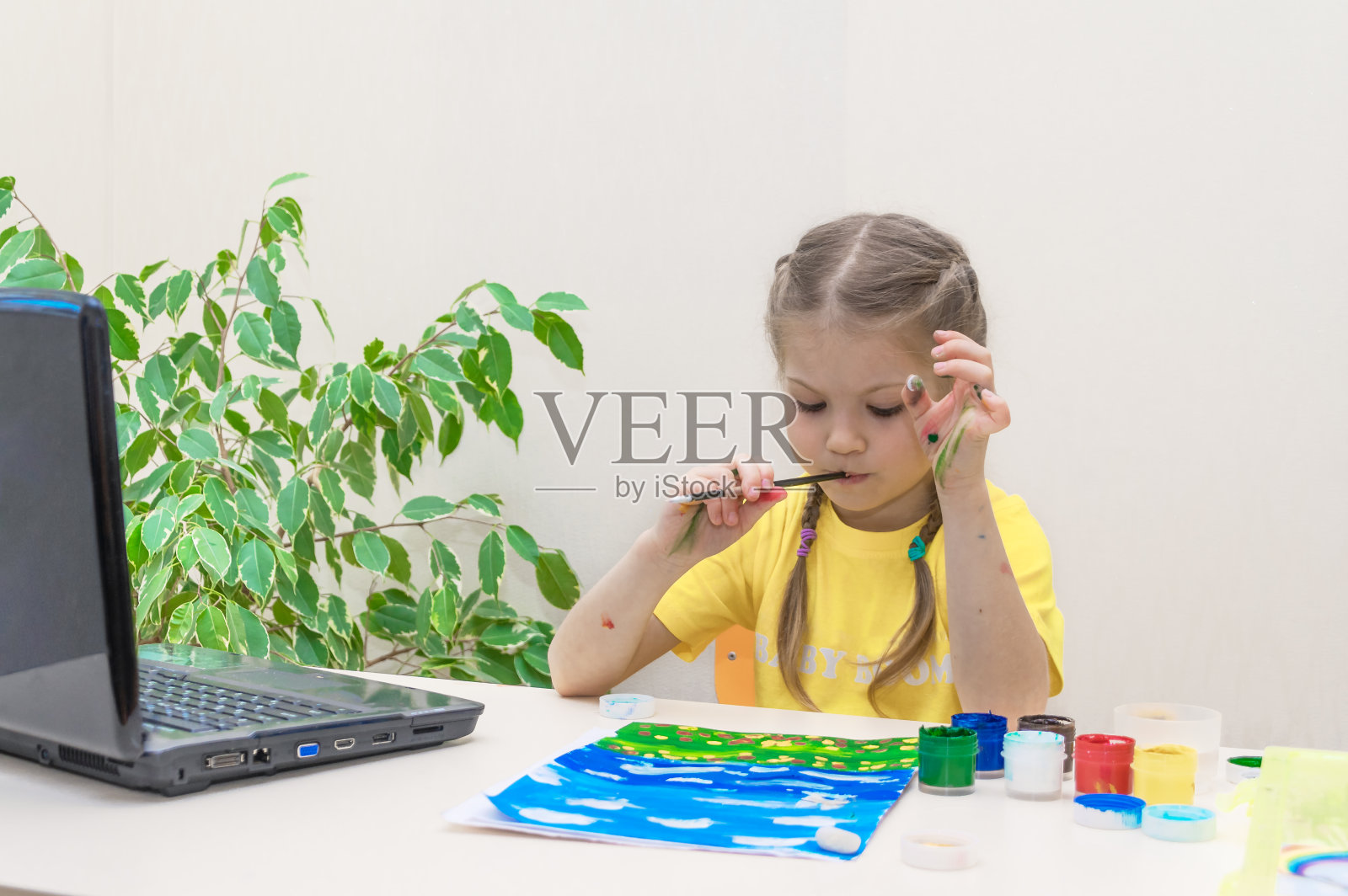 远程学习在线教育。一个学龄前的女孩在家里学习画画，看她的笔记本电脑，做她的家庭作业。女孩的想法。刷在嘴里。画水粉，在桌子上画画。照片摄影图片