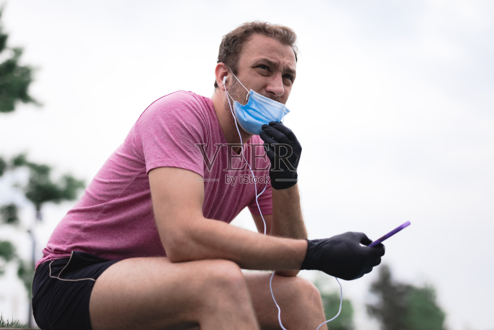 运动员戴着医用口罩和手套，智能手机和耳塞，在城市环境中慢跑。精疲力竭的男子从慢跑和医用口罩的使用。麻烦与呼吸。照片摄影图片