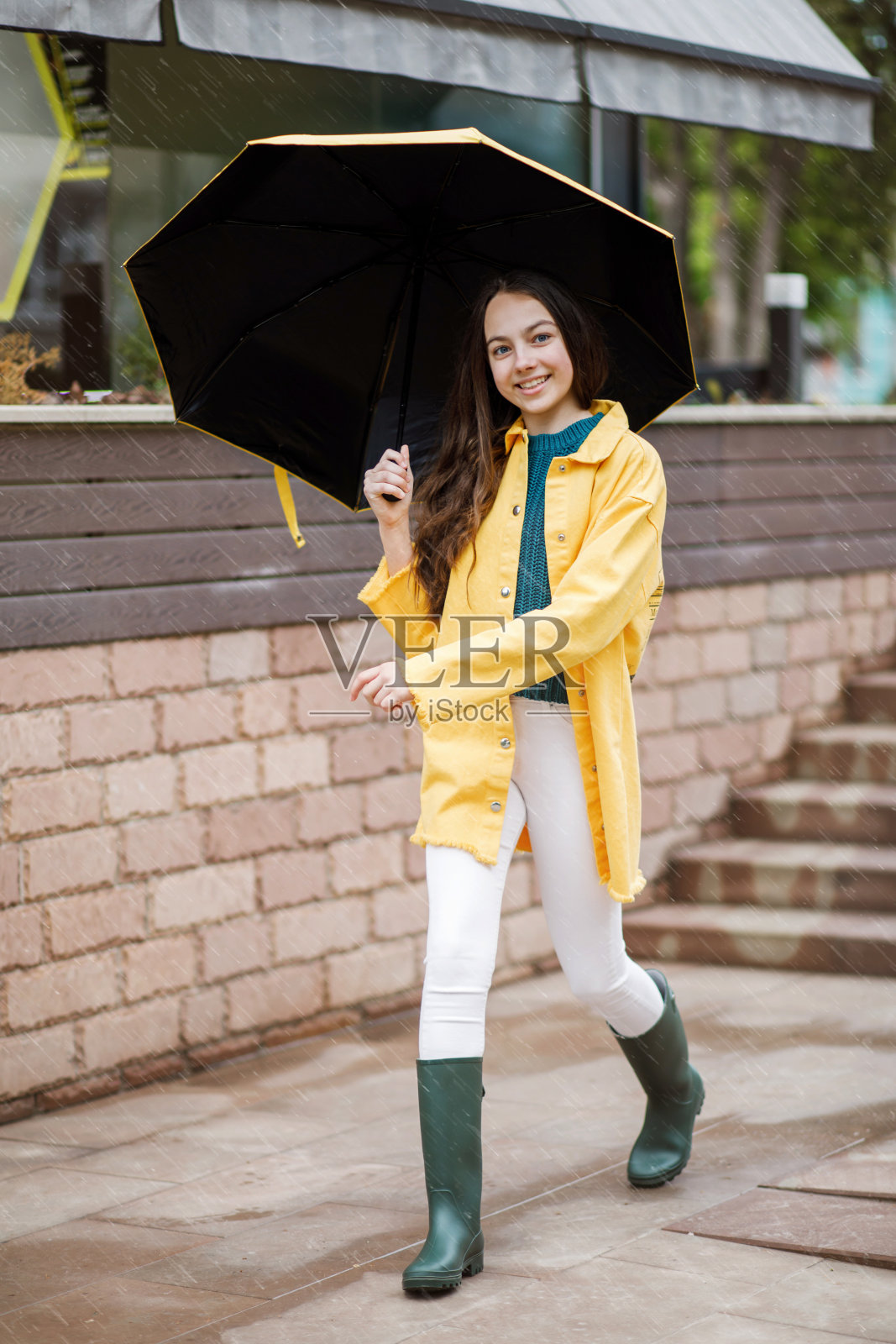 一个年轻漂亮的少女走在城市的雨天。这个女孩穿着彩色的衣服和胶靴照片摄影图片