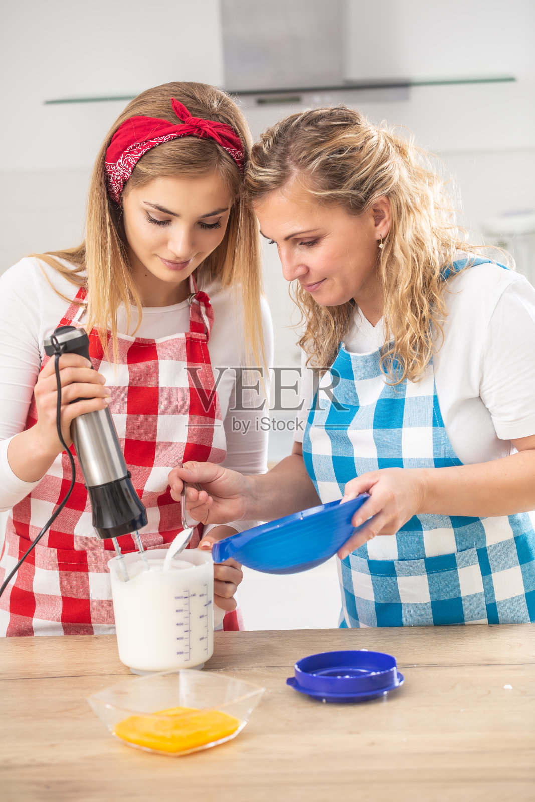 金发碧眼的母亲穿着格子围裙在厨房里帮助女儿烘焙。照片摄影图片