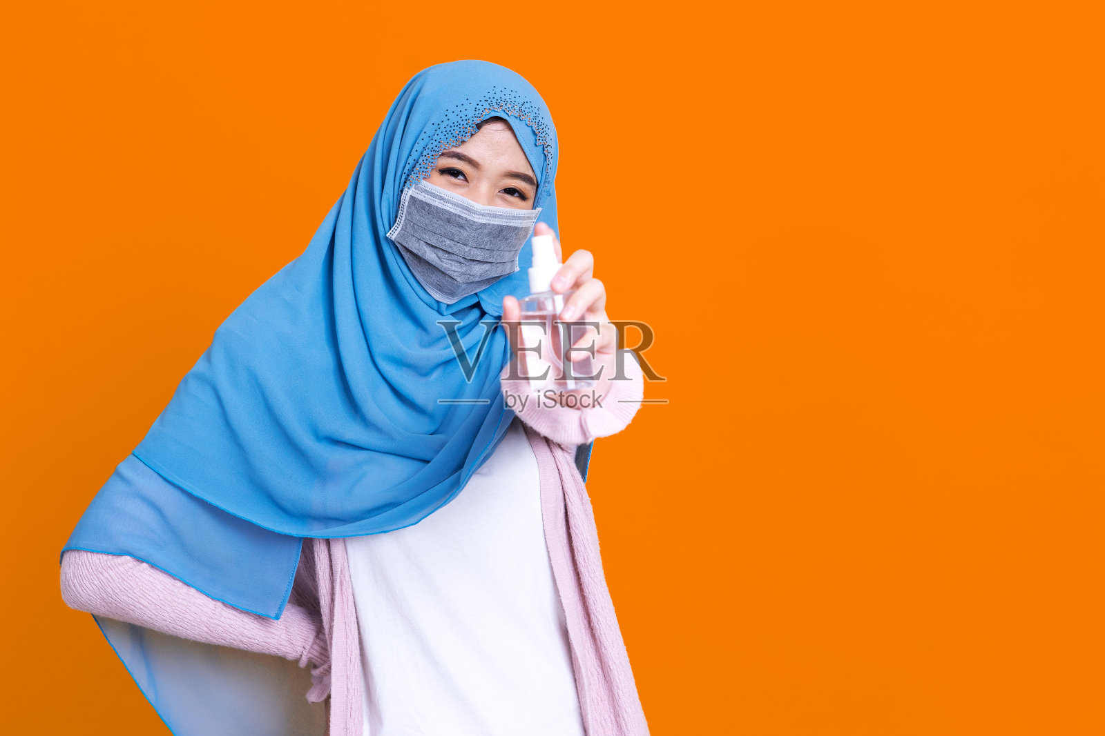 在病毒爆发时，穿着传统服装的亚洲穆斯林妇女戴着医用口罩，用分离的彩色背景酒精凝胶洗手照片摄影图片