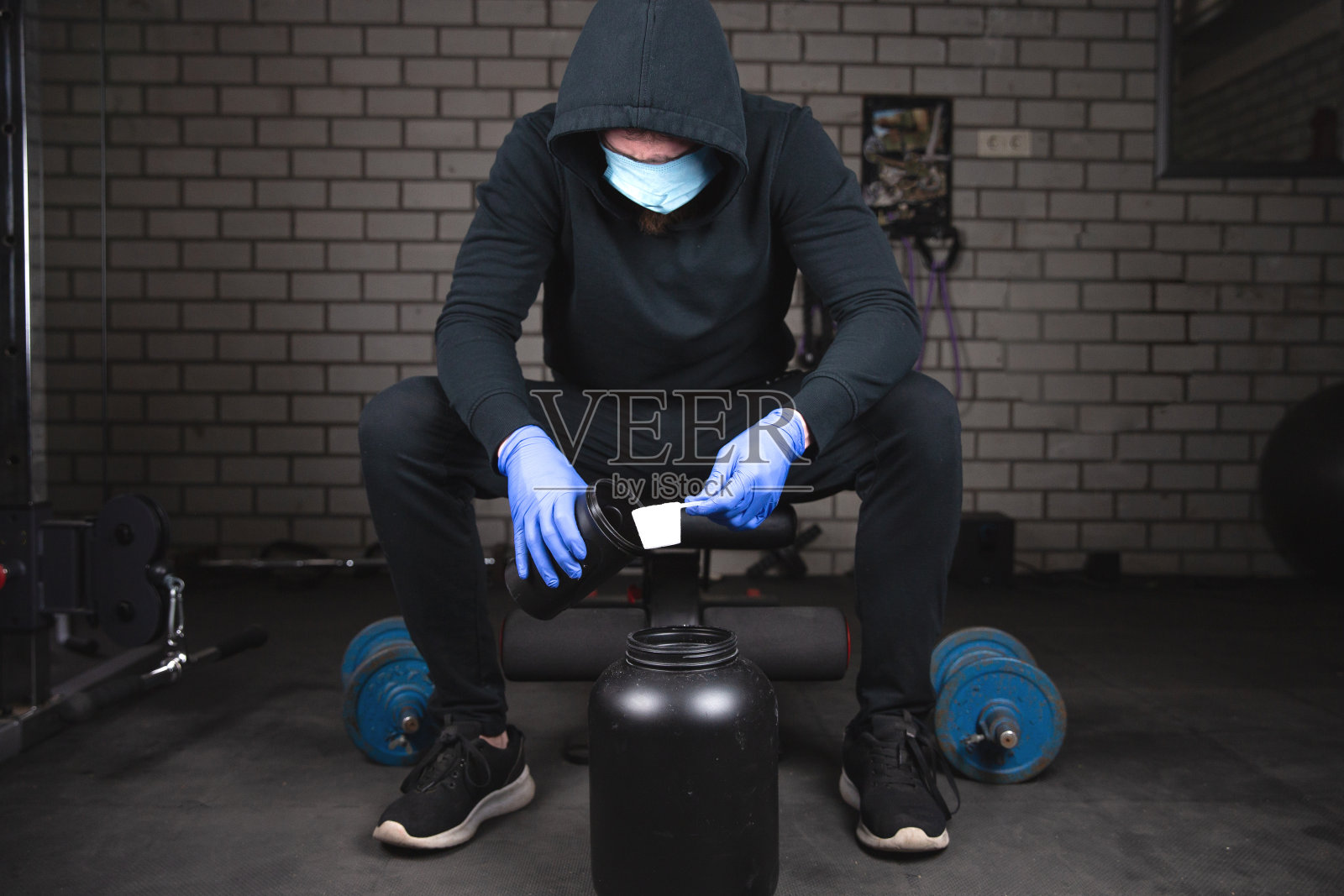 一名健康青年在健身房准备蛋白质奶昔，以更快地锻炼肌肉，他穿着黑色运动服，戴着防护口罩和手套，以应对新冠病毒照片摄影图片