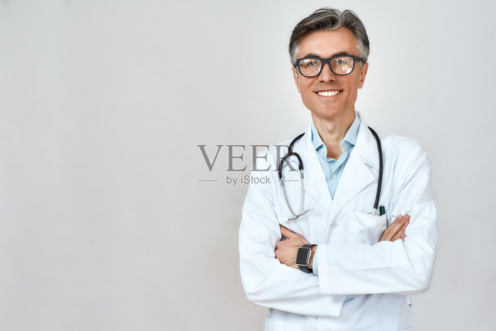 快乐的资深男医生，穿着白大褂，脖子上挂着听诊器，双臂交叉，对着镜头微笑，站在灰色的背景下照片摄影图片