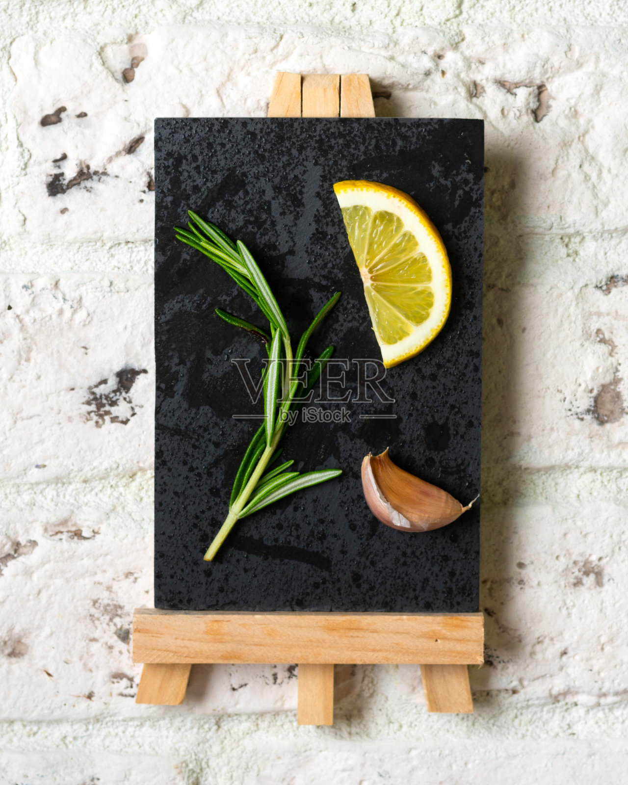 一枝迷迭香，一片柠檬和一瓣大蒜放在一块石板上，背景是白色的砖块。图片、明信片或日历的概念照片摄影图片