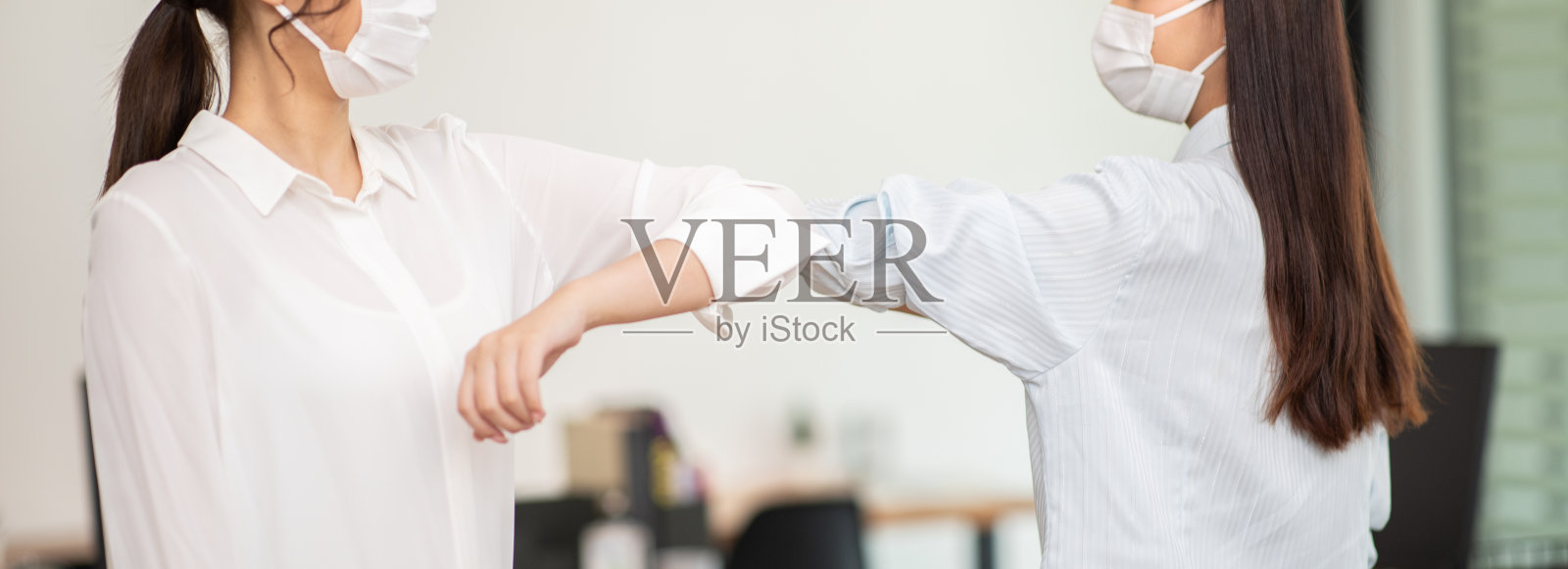 由于COVID-19大流行，两名女商人戴着防护口罩，在工作场所冲撞肘部以保护病毒照片摄影图片