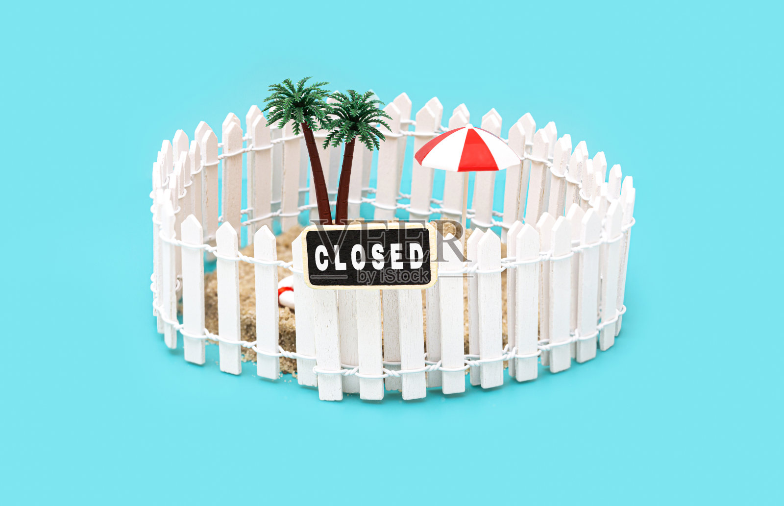 用栅栏围起来的微型玩具岛，有棕榈树，太阳伞和一个写着“在浅蓝色的背景上关闭”的标志。照片摄影图片
