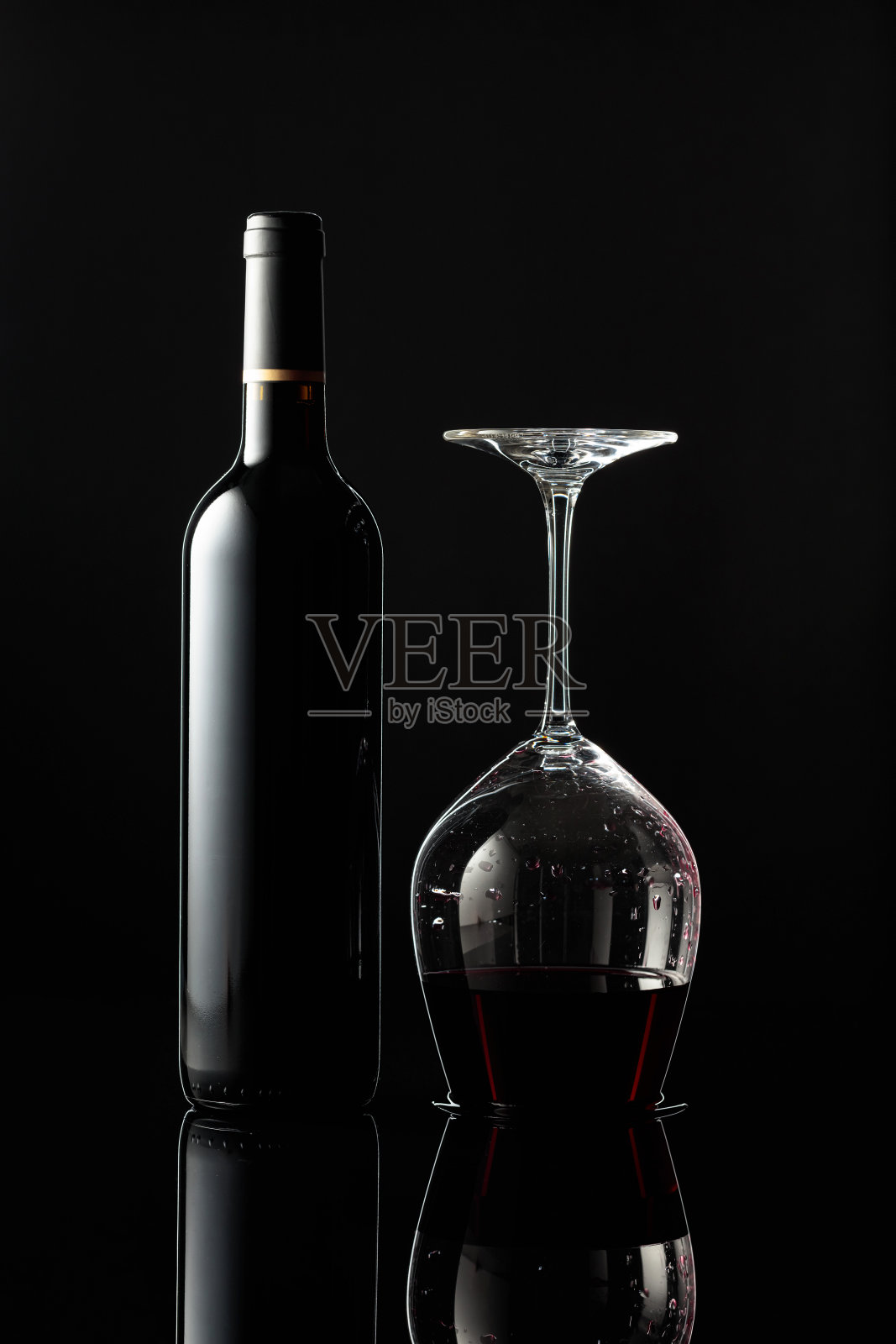 一瓶红酒和一个倒置的杯子，葡萄酒在一个黑色的背景。照片摄影图片