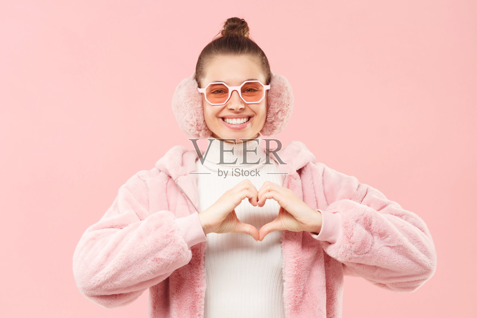 年轻的女孩穿着冬天的大衣，戴着有色眼镜和耳罩，用手表示爱心，分享爱和欣赏，孤立在粉红色的背景下照片摄影图片