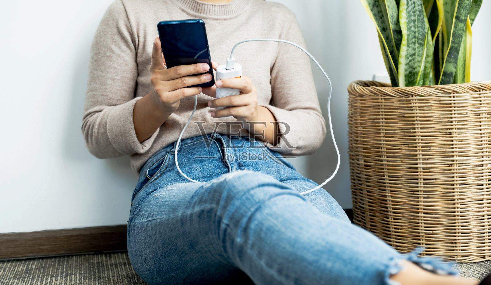一名年轻女子靠墙坐着，手里拿着一部智能手机，正在给智能手机充电照片摄影图片