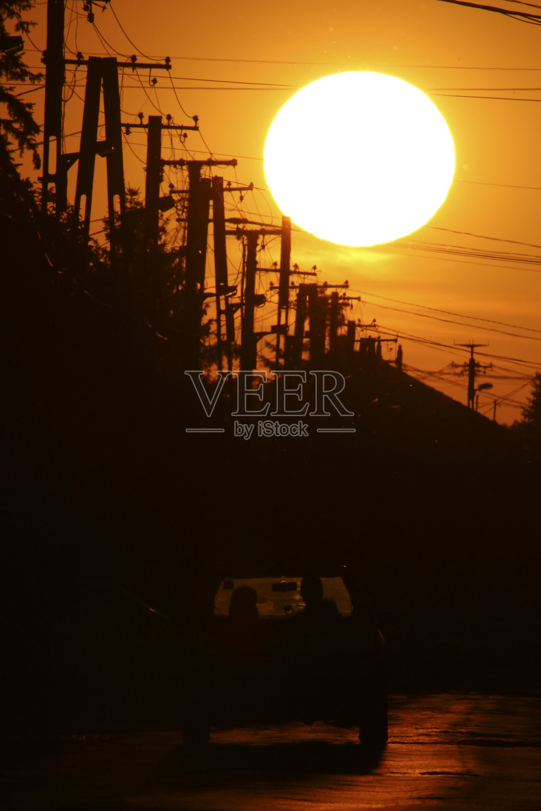 日落时分，在通往城市的道路旁可以看到电网，一辆汽车和两名乘客的剪影。天空是橙色的，太阳很大。照片摄影图片