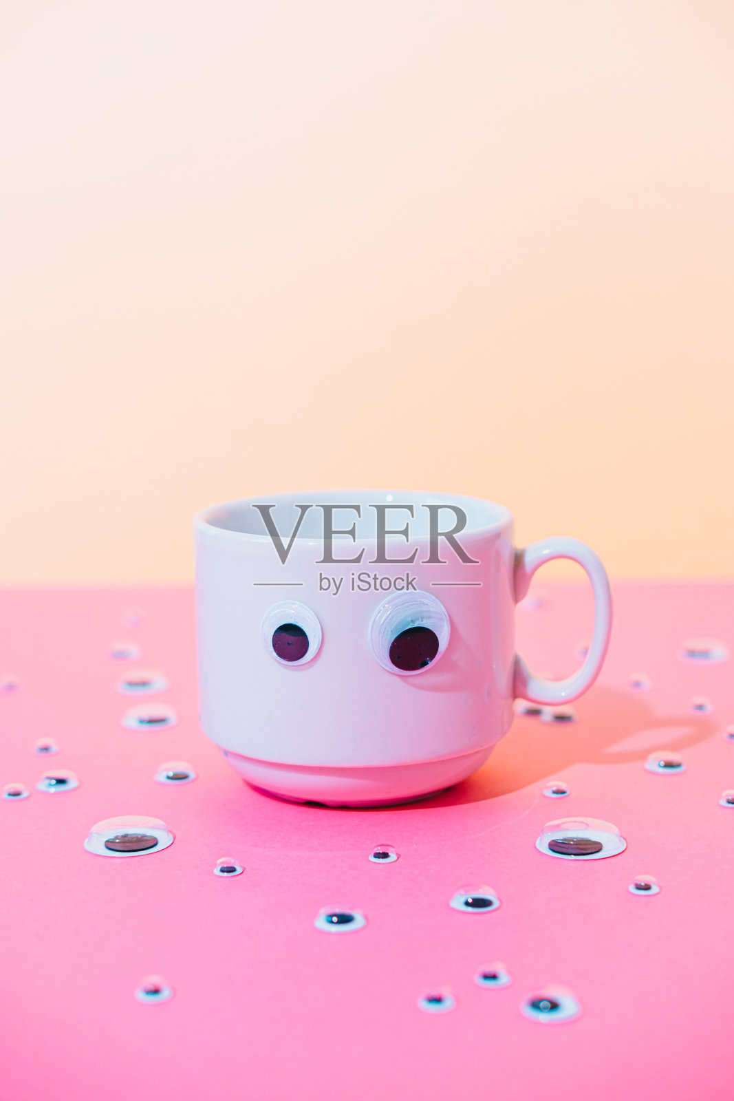 一杯热咖啡或茶与卡通玩具眼睛在粉红色和黄色柔和的超现实主义背景。照片摄影图片