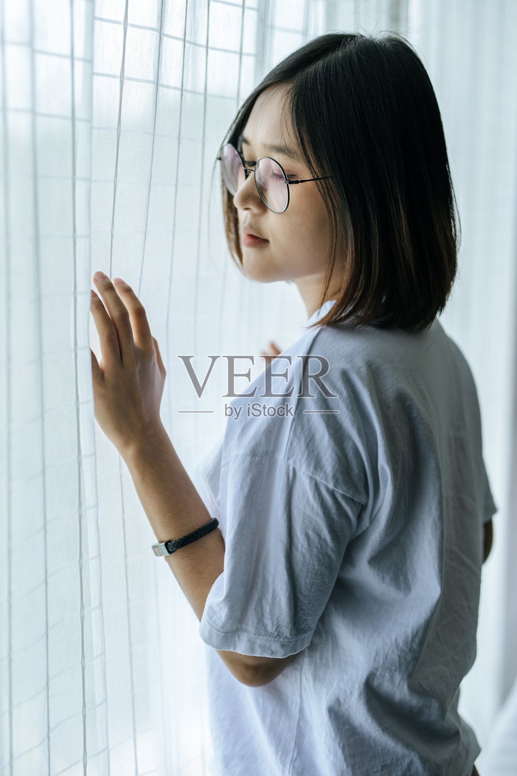 一个穿着白衬衫的女人，站在窗前，抓住窗帘。照片摄影图片