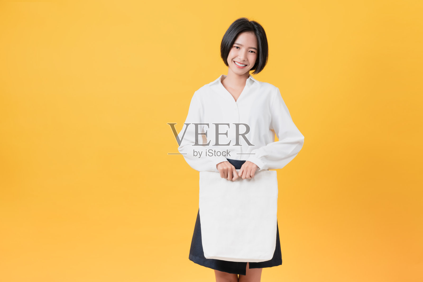 微笑的亚洲女人在休闲白色t恤站和拿包帆布织物的样品标志在黄色的背景。照片摄影图片