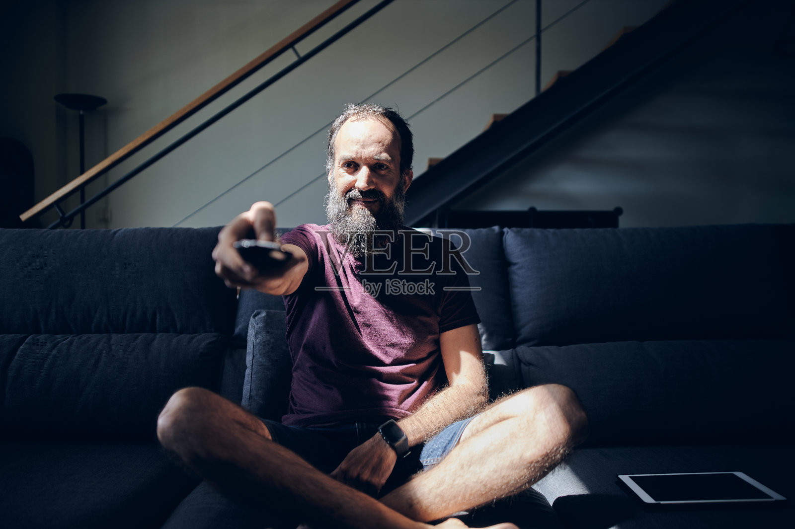 满脸胡须的男子微笑着坐在沙发上，旁边放着一台平板电脑，用遥控器打开电视照片摄影图片