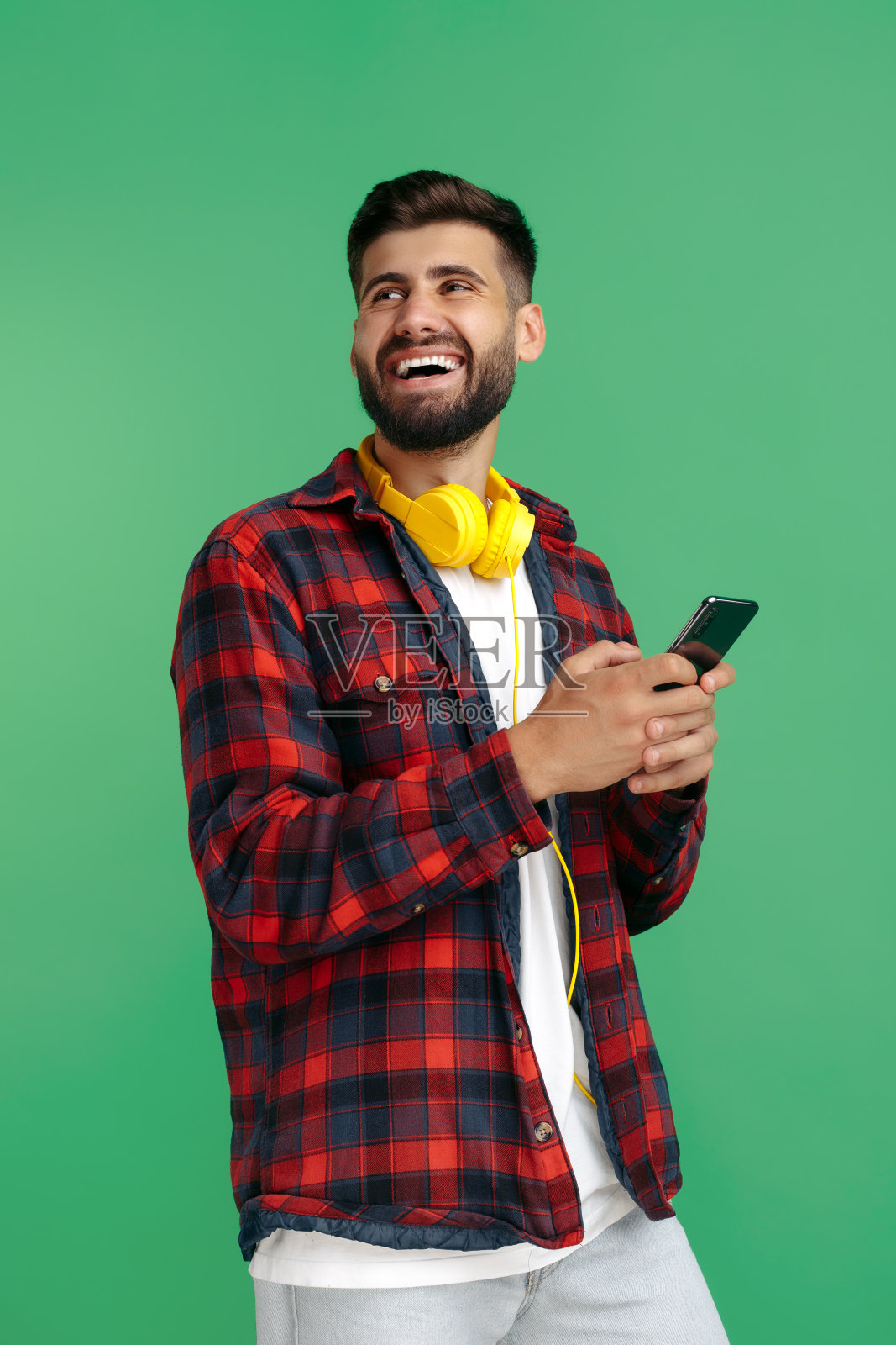 一名快乐的留着胡子的潮男穿着格子衬衫，戴着耳机在绿色背景上使用智能手机。照片摄影图片