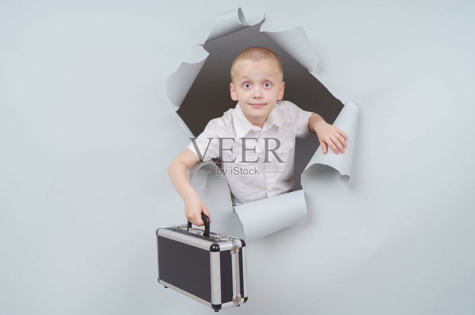 酷男孩提着一个装满钱的手提箱从工作室的破纸洞里冒了出来，带着兴奋愉悦的表情，透过灰色的背景看着。照片摄影图片