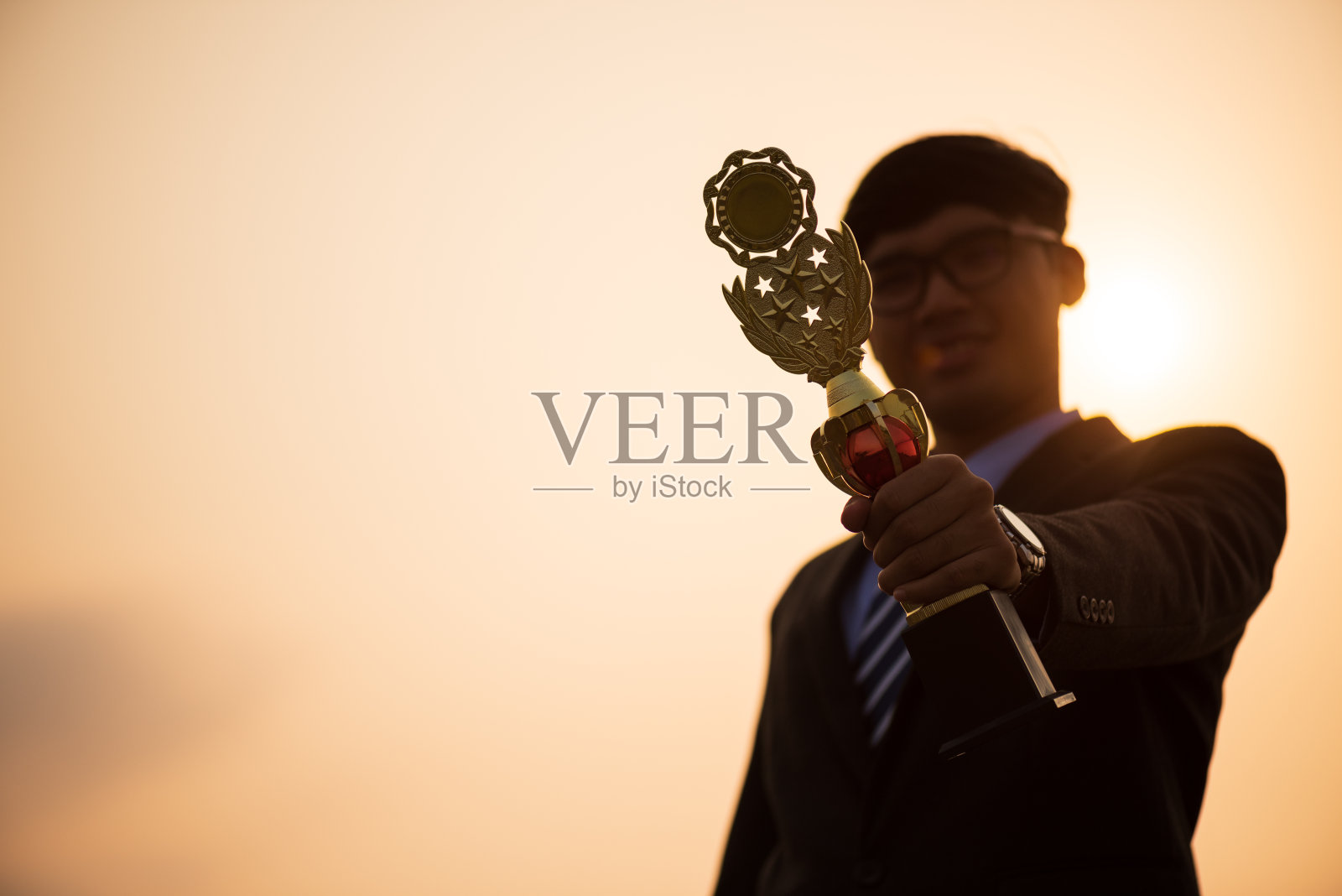 一个快乐的亚洲英俊的年轻商人在商业套装显示金像奖获奖奖杯在日落日出山的背景。经营理念。照片摄影图片