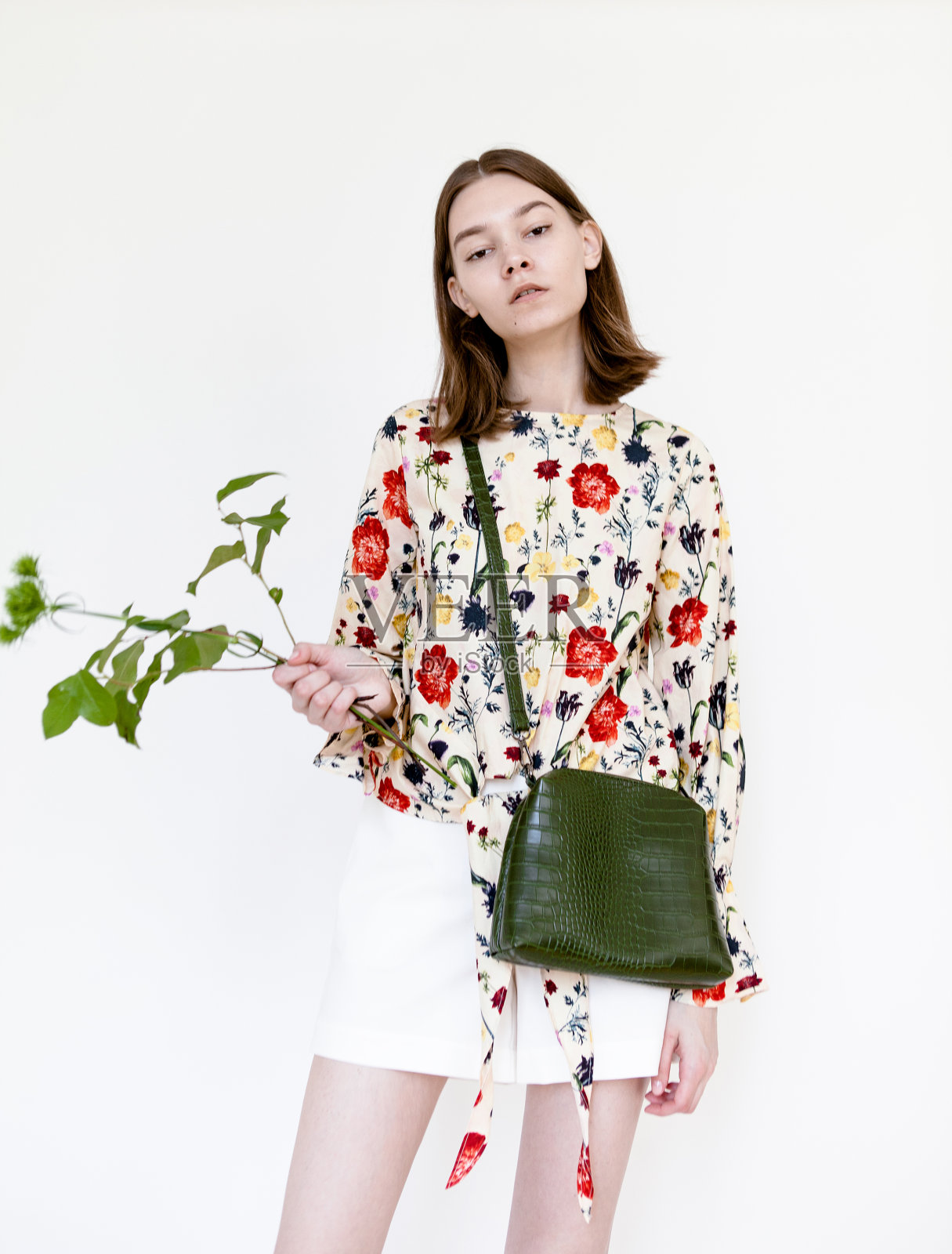 美丽的年轻女子与裸色化妆穿花印花衬衫，白色裙子和黑色包拿着一朵花在白色的背景照片摄影图片