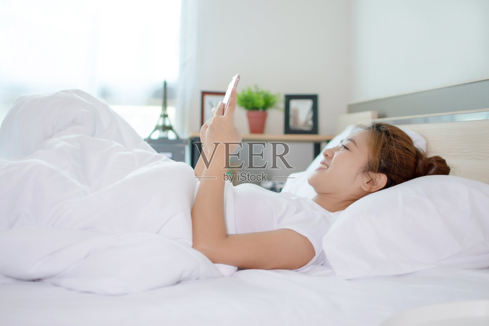 一名微笑的亚洲女子正在使用智能手机，她睡在卧室的床上。照片摄影图片