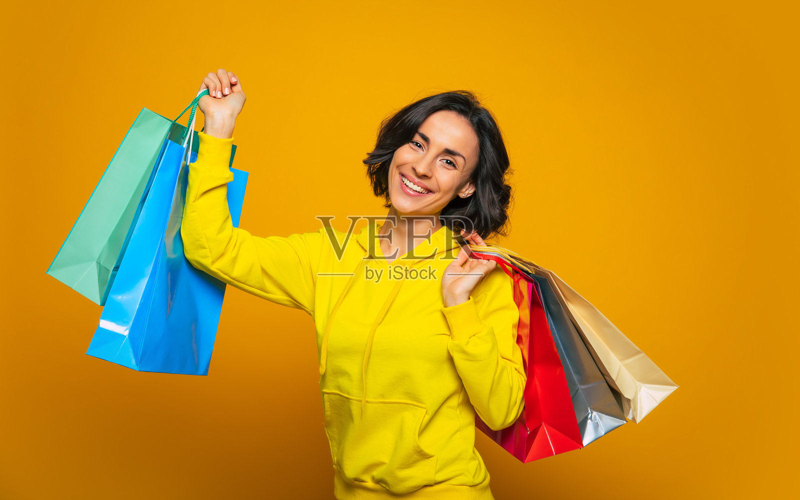 我终于得到了我想要的!一个年轻女子的半长照片，穿着黄色的帽衫，因为购物而高兴，拿着她在彩色的袋子里买的东西。照片摄影图片