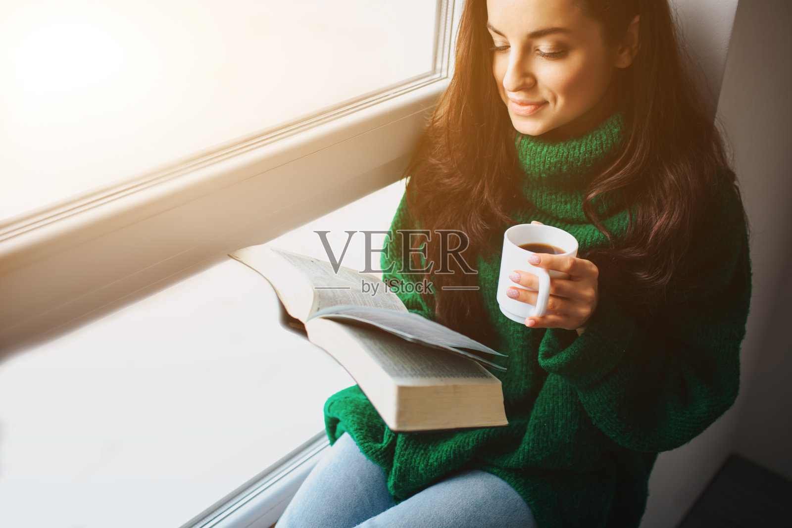 完美的早晨一个年轻的黑发女子坐在窗台上，手里拿着一本书和一杯茶或咖啡。女模特穿着绿色的宽松毛衣。照片摄影图片