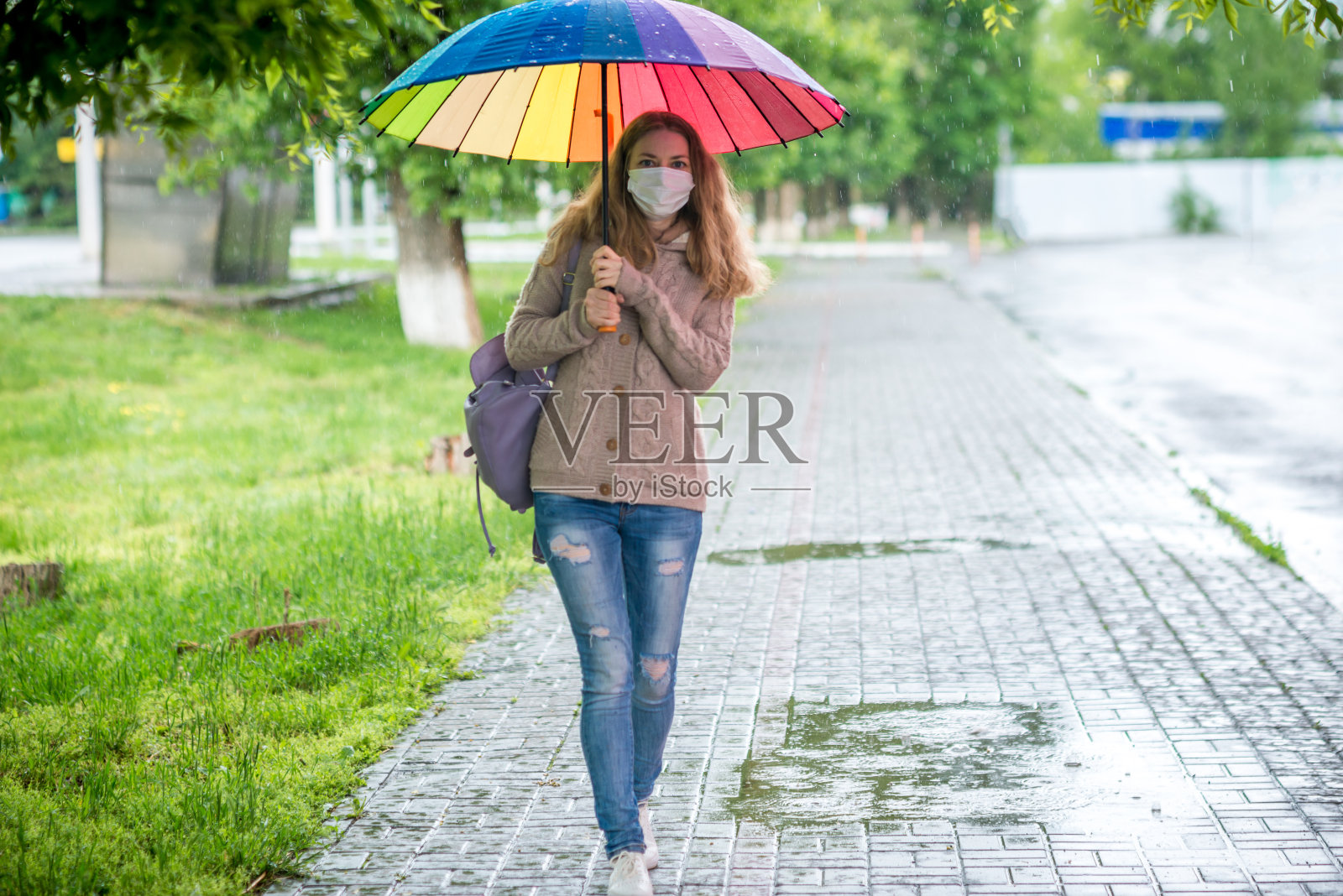 在春雨中，一个戴着保护面罩的白人女孩在雨伞下走在空旷的街道上。冠状病毒大流行期间的安全和社会距离。新常态，隔离的含义。照片摄影图片