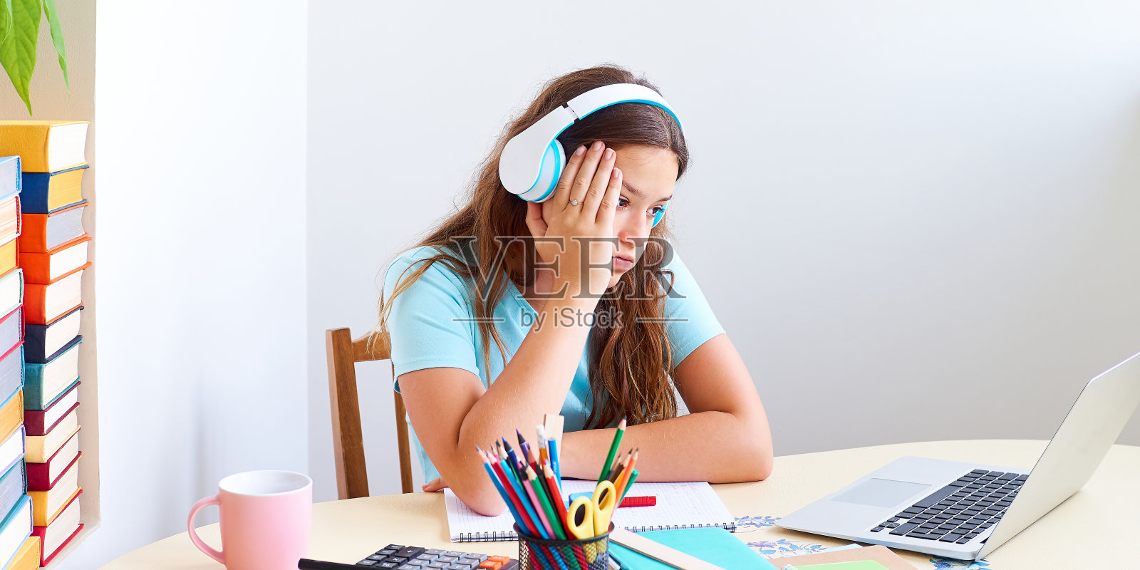 戴着耳机的少女学生坐在桌子前看着笔记本电脑屏幕，厌倦了学习。照片摄影图片