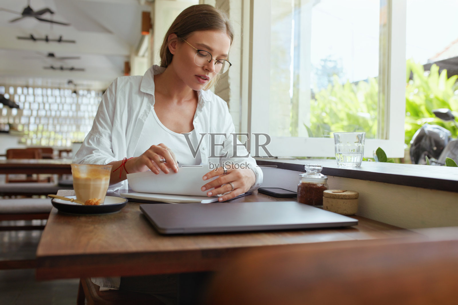 业务。在咖啡馆远程工作的女人。穿着休闲装戴着眼镜的女孩用笔记本电脑在笔记本上写东西。数字技术为暑期在舒适的地方创造性的工作。照片摄影图片