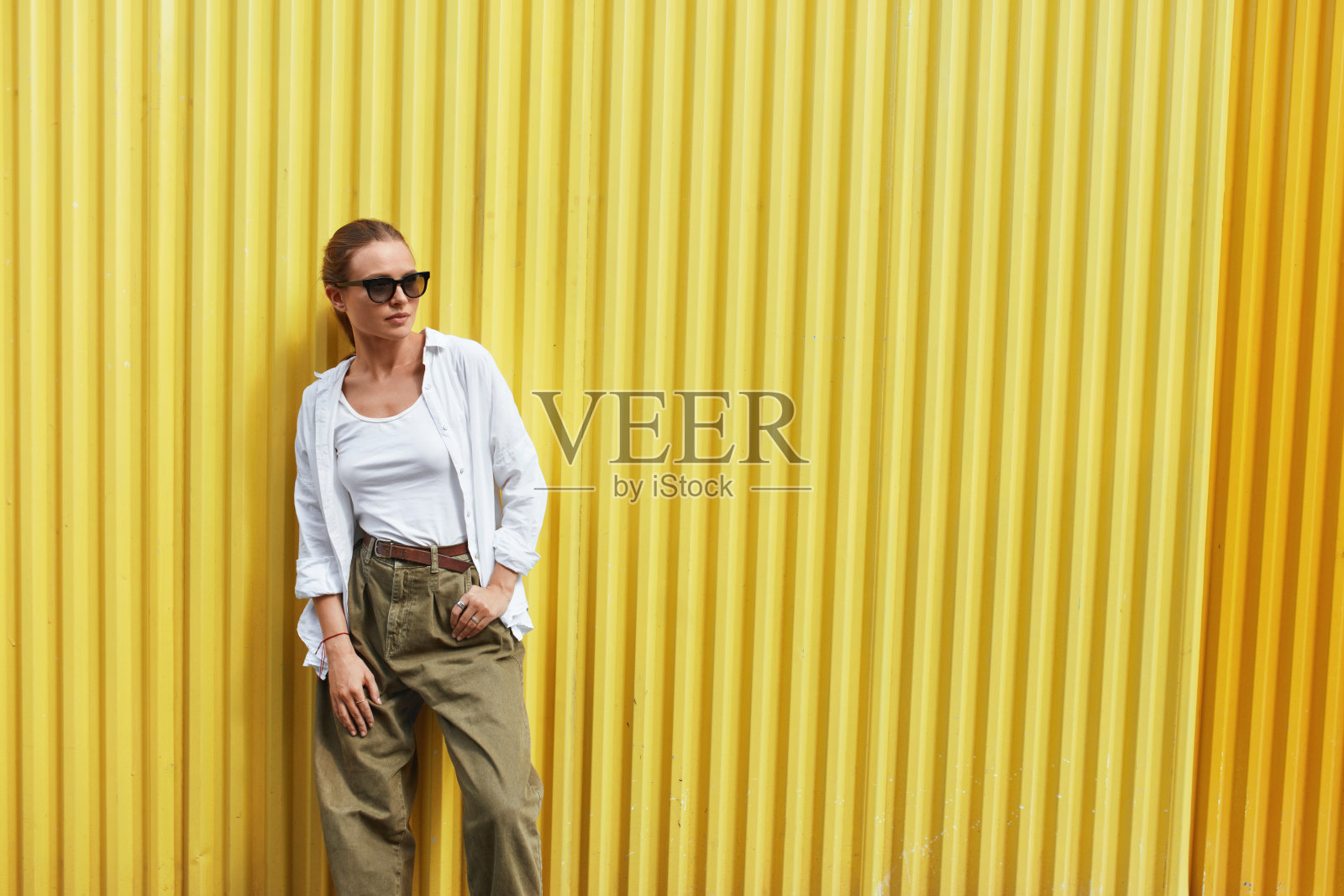 时尚的女孩。女人的肖像。时尚模特穿着休闲服，戴着太阳镜，站在黄色的金属栅栏前。女性在白色衬衫和背心。城市的生活方式。照片摄影图片