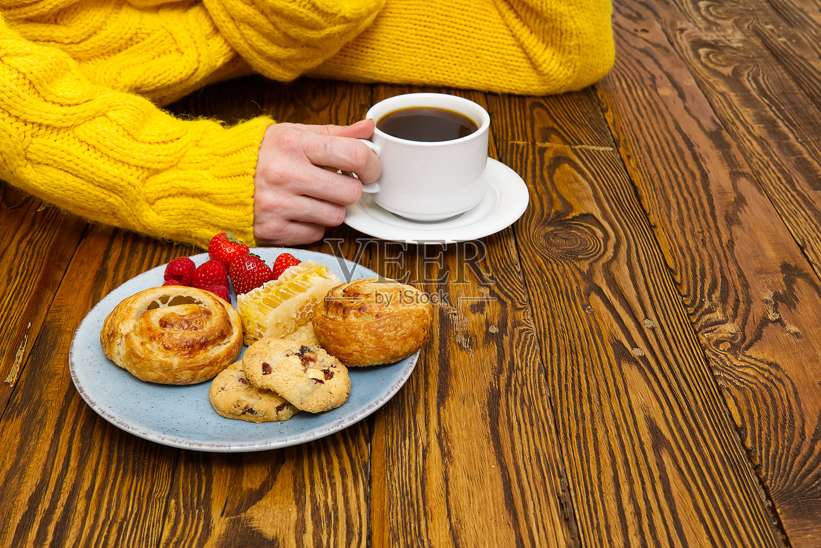 穿着黄色毛衣的女性双手捧着咖啡和饼干放在棕色的旧木桌上。照片摄影图片