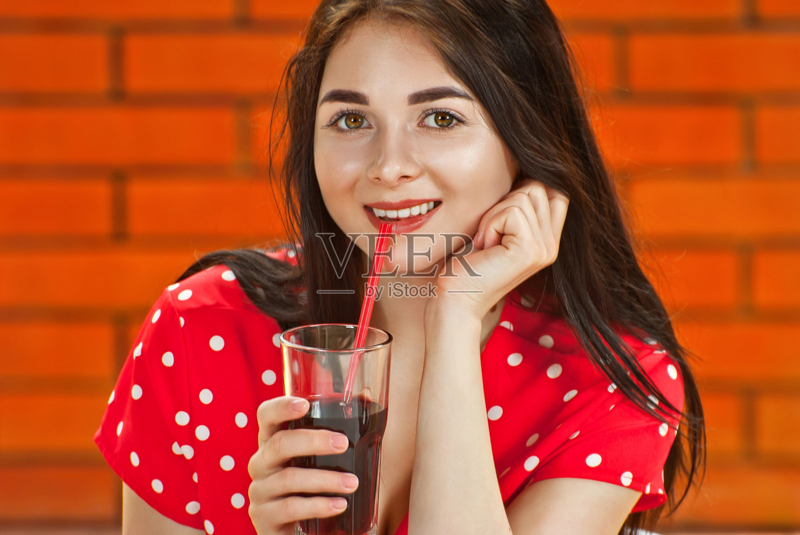 一个快乐的女孩坐在街边咖啡馆的桌子旁。一个穿着红色白色圆点连衣裙的女人喝果汁。橙色的砖墙在背景中模糊了。照片摄影图片