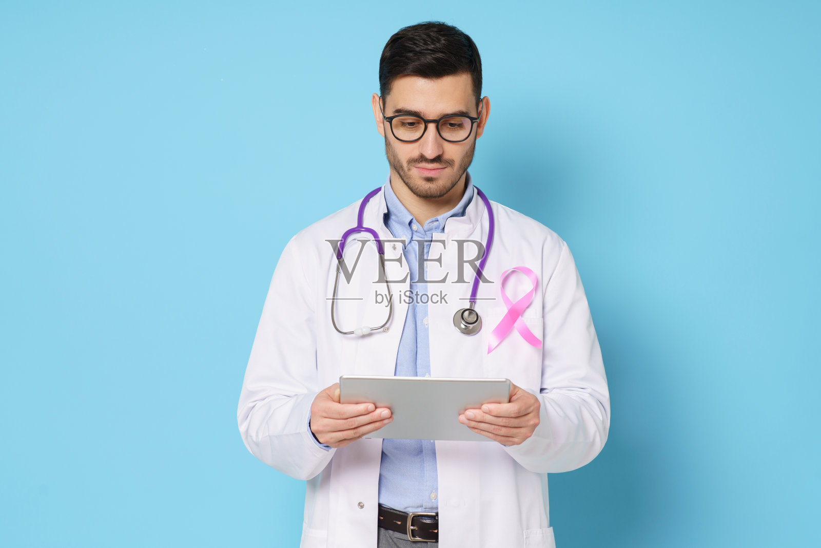 年轻的男性肿瘤医生拿着平板电脑，专注地看着屏幕，他的长袍上系着粉色的丝带，在蓝色的背景下，他感觉自己很负责，很孤独照片摄影图片