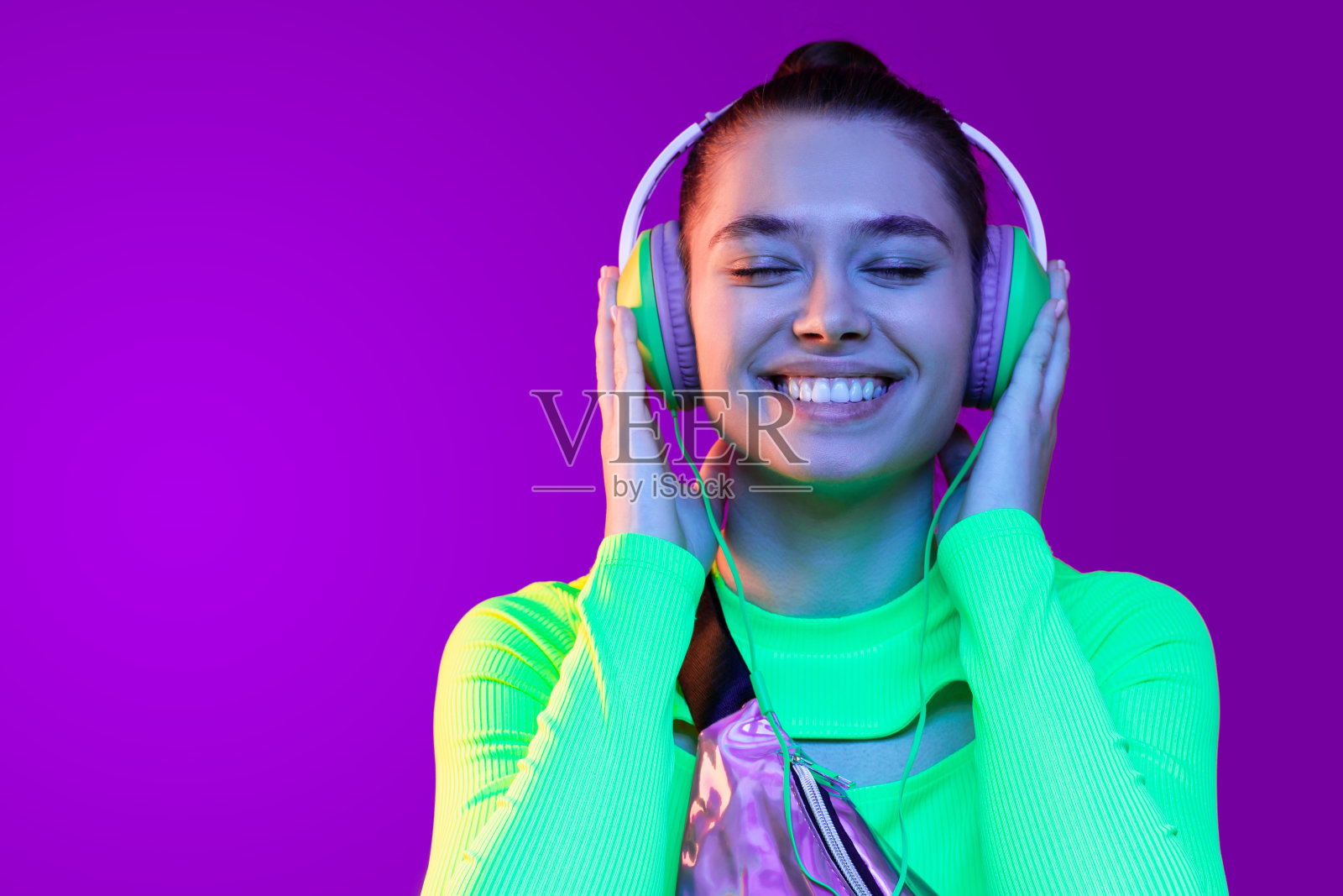 年轻快乐的女孩站在绿色的霓虹顶，戴着耳机，享受喜爱的音乐，闭着眼睛，孤立在紫色的背景照片摄影图片