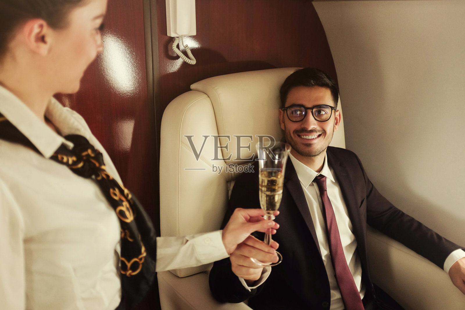 一位年轻的商人坐头等舱，穿着西装坐在桌边，空姐正给他端来一杯香槟庆祝他的成功照片摄影图片