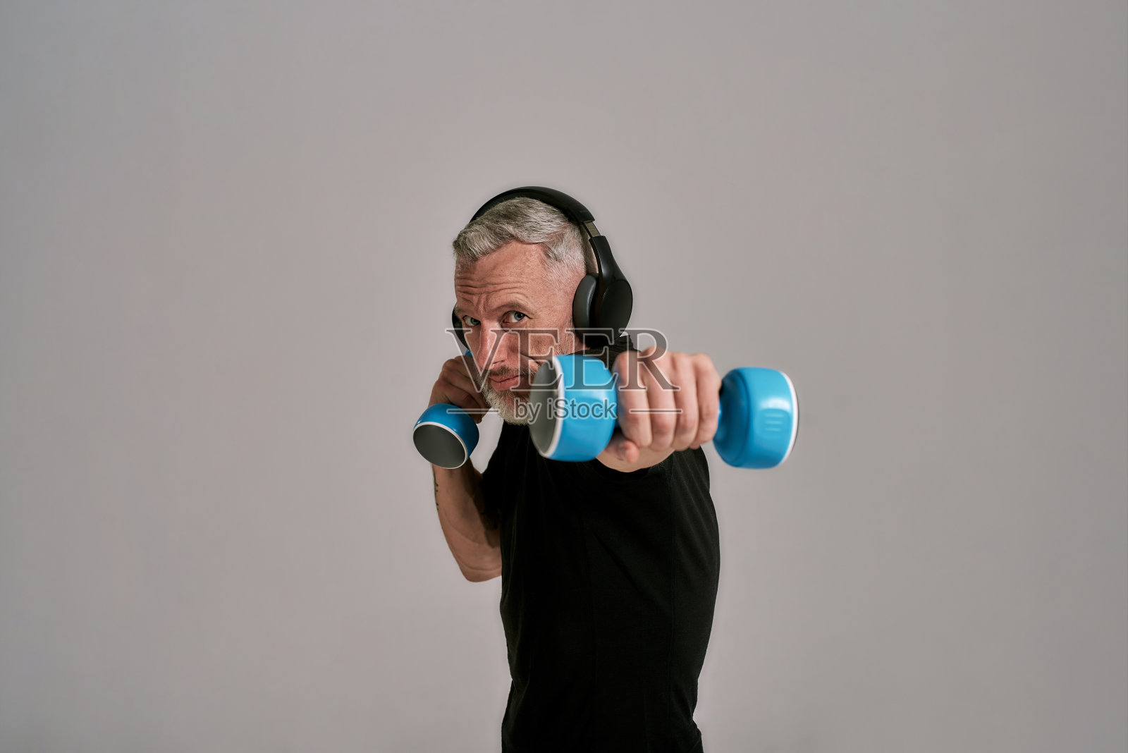 中年肌肉男穿着黑色t恤，戴着耳机，拿着蓝色哑铃看着摄像机，在工作室灰色的背景下摆姿势照片摄影图片