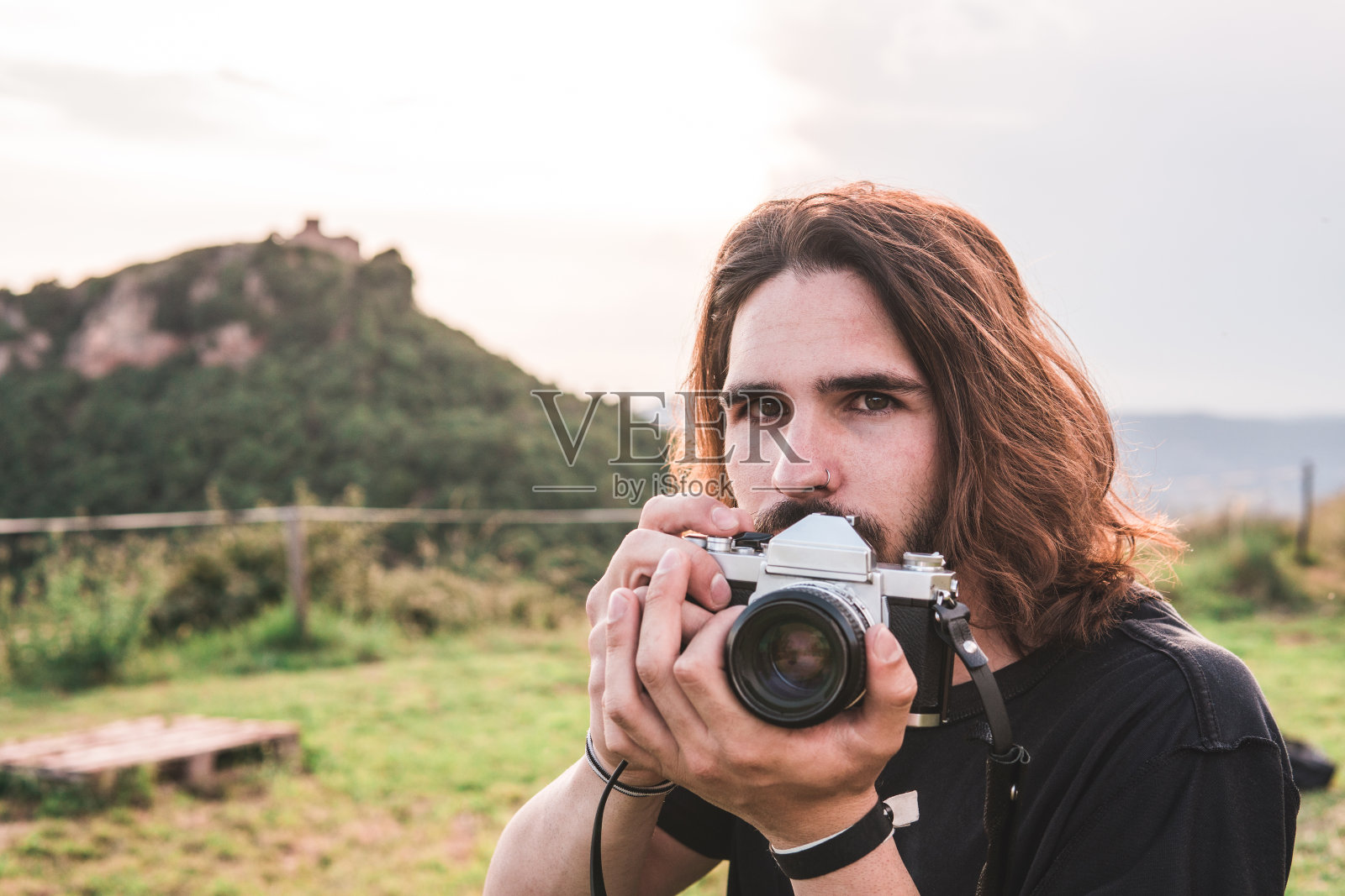 一个喜欢冒险的年轻人看着镜头。他在看他的相机。他身后是一座山，山顶上有一座建筑物。照片摄影图片