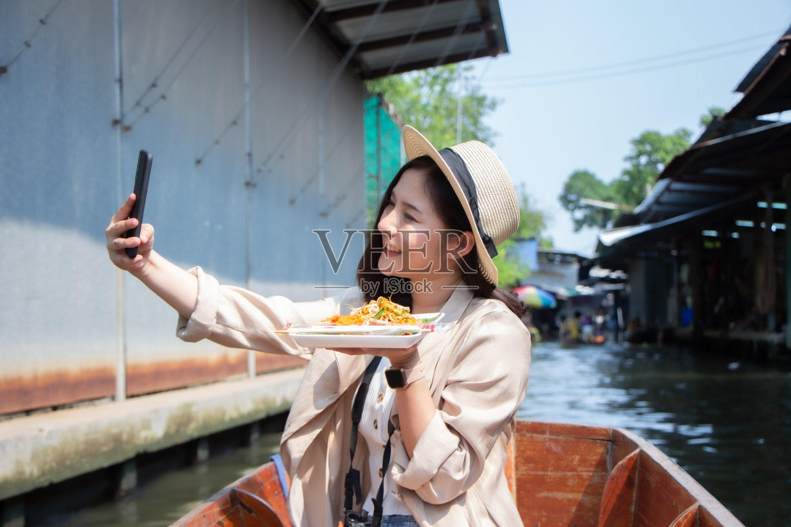 亚洲女性游客戴着帽子拿着相机和智能手机拍摄视频直播。美丽的女人乘坐出租船访问达姆努恩Saduak浮动市场，Ratchaburi。照片摄影图片