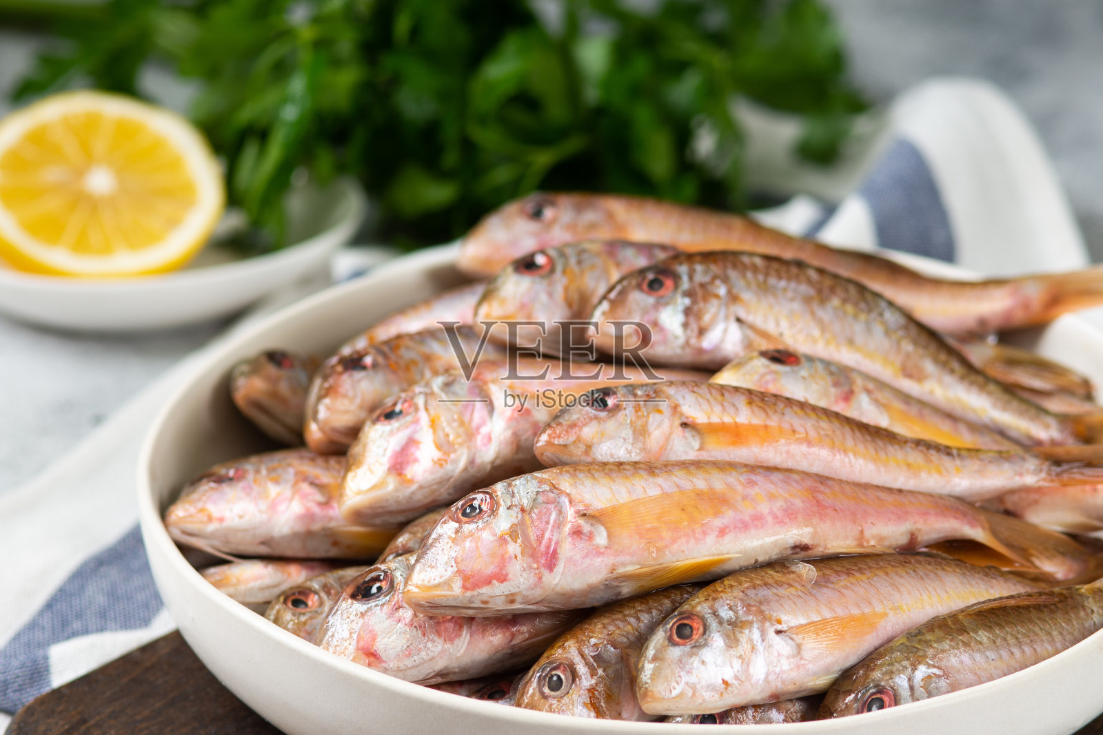 在浅灰色的餐桌上，白色的陶瓷碗里放着黑色的海鱼。很多鱼，红鲻鱼。黑海barabulka照片摄影图片