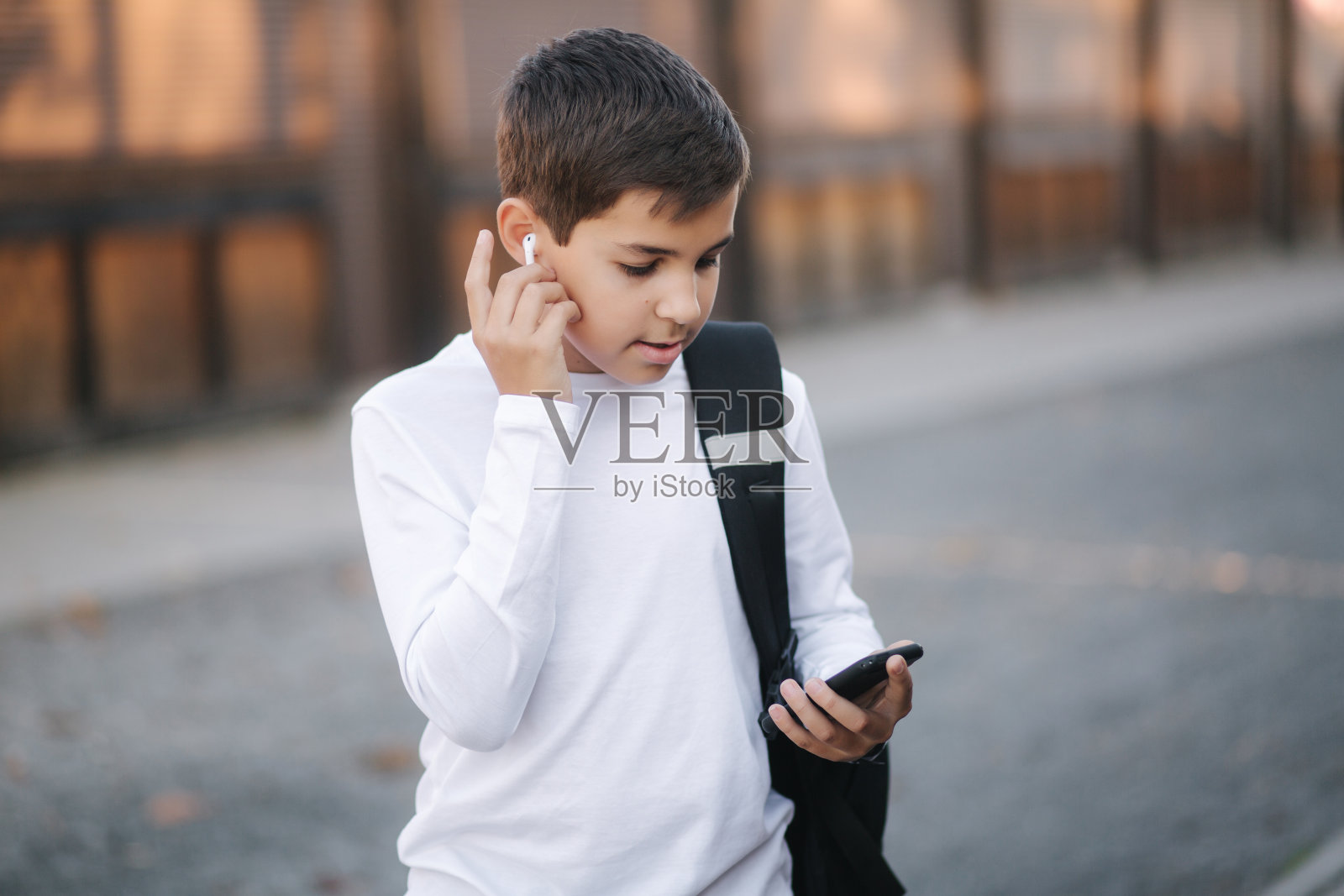 十几岁的男孩拿出一个无线耳机，然后放进耳朵里。穿着白衬衫背着背包的小男孩。男孩听音乐照片摄影图片