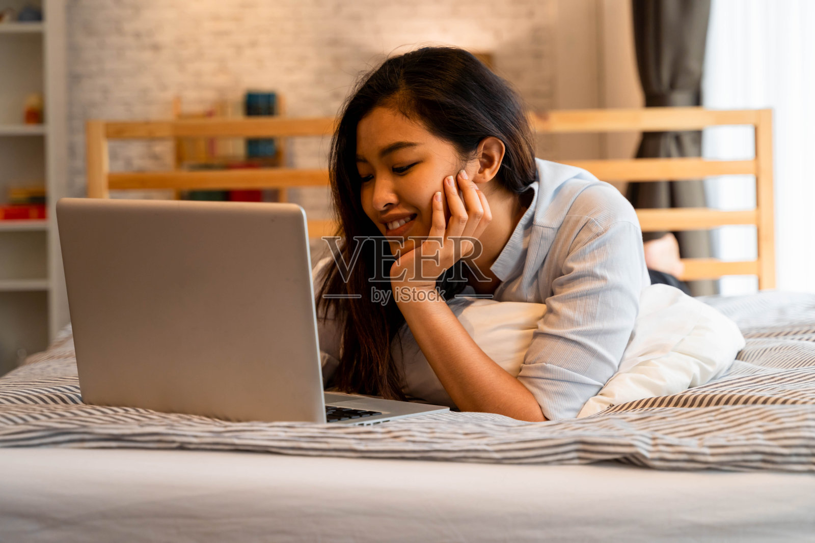 快乐的亚洲女人在家里的床上用笔记本电脑工作。随意美丽的女孩在家里微笑，而工作在家照片摄影图片